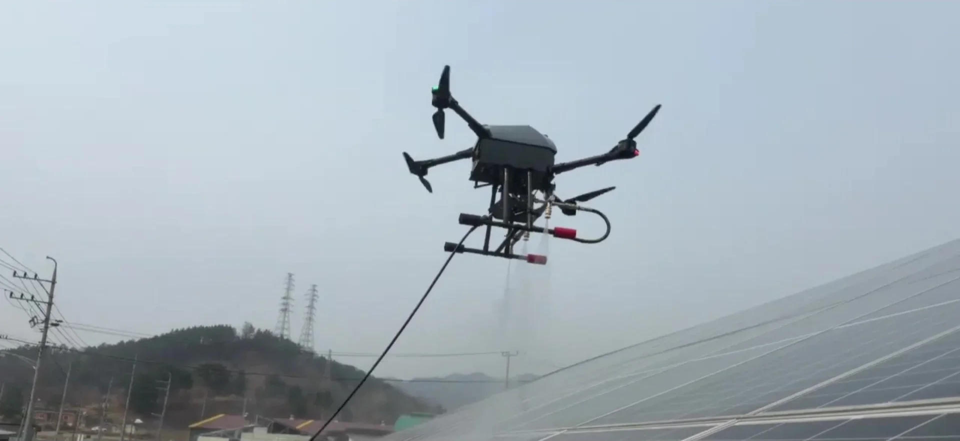 지상에서 조종하는 드론이 패널 위에서 물을 뿌리고 있습니다. 호스가 연결된 상태로 비행합니다.
